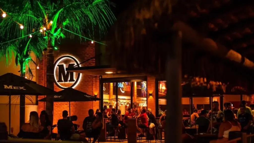 Exterior iluminado do Mex Guarapari à noite, na Praia do Morro, com o logo "M" brilhando e clientes em mesas. Fim De Semana Em Guarapari: Festival Em Setiba E Baladas No Mex