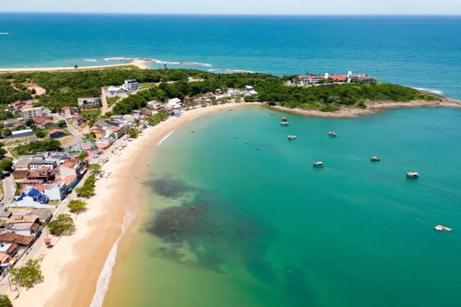 Vista aérea da curva da Praia de Ubu em Anchieta, Espírito Santo, mostrando o mar verde cristalino e a orla com casas e árvores.