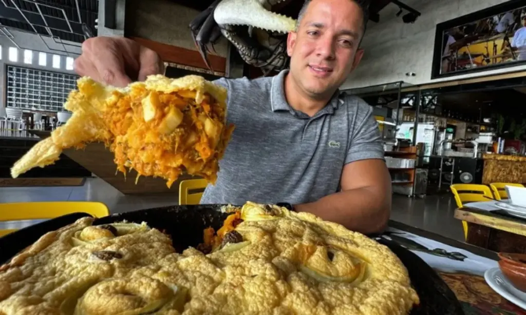 Homem sorridente em um restaurante servindo uma fatia generosa de Torta Capixaba retirada de uma panela de barro, durante o Festival Nos Passos da Torta Capixaba.