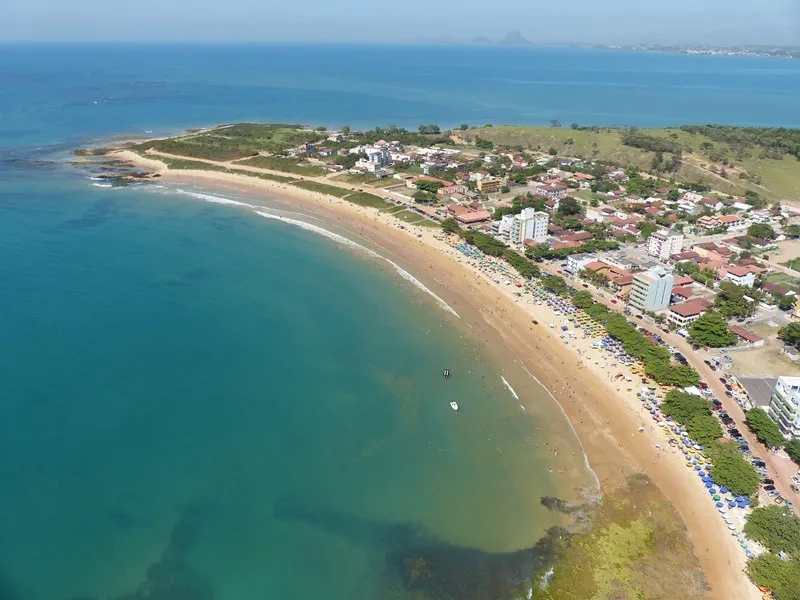 Vista aérea da orla do Balneário de Castelhanos em Anchieta, mostrando a extensa faixa de areia clara, águas calmas e vegetação de restinga preservada.